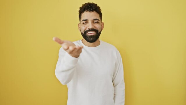 Cheerful young man offers a friendly palm, standing isolated on a yellow background, signifying acceptance and giving assistance