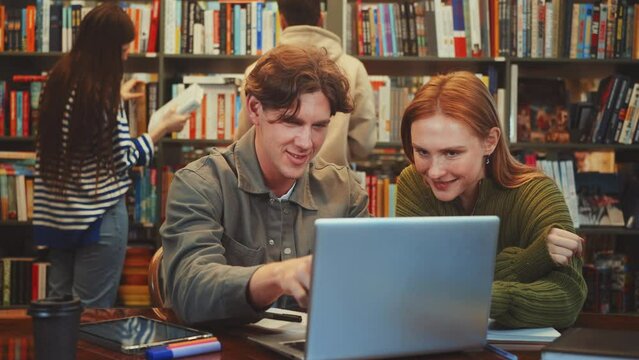 Two focused students engage in collaborative study using laptop at library table, surrounded by shelves packed with colorful books. Another persons browse books in background