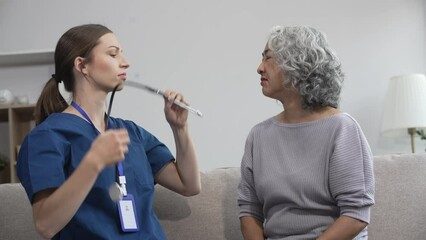 Caucasian female doctor uses stethoscope to check heart rate on elderly Asian patient on sofa.