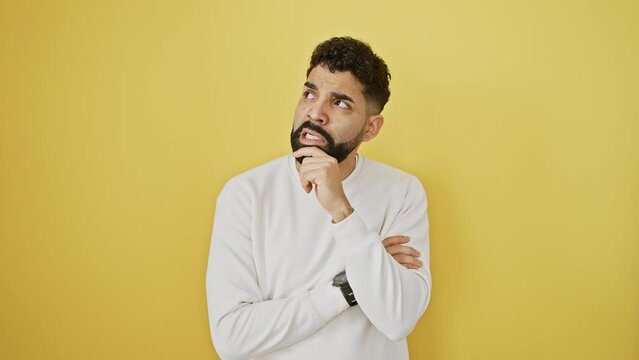 Worried young man, hand nervously on chin, standing troubled, thinking about his complex question on a yellow isolated wall.