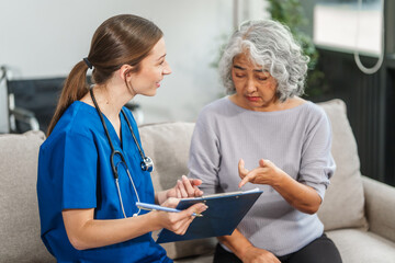 Caucasian teenager female doctor consults with and Asian elderly patient on medical history, discussing headaches, chest pain, coughing, sneezing, stomach pain, and neck pain while seated on a sofa.