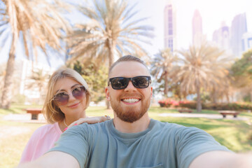 Fototapeta premium Selfie photo of happy couple young man and woman tourists background park with palm and skyscrapers in Dubai, UAE tourism travel