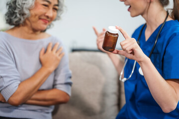 Caucasian female doctor introduces medicine to an elderly Asian patient while seated on a sofa, providing guidance and information about the prescribed medication.