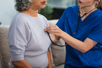 Caucasian female doctor uses stethoscope to check heart rate on elderly Asian patient on sofa.