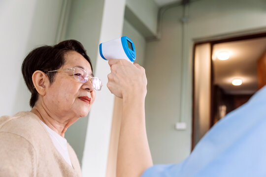 Woman nurse measures the temperature of an elderly Asian woman by using an infrared forehead thermometer gun at her home. Caregiver visit at home. Home health care and nursing home concept.