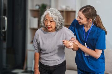 Obraz premium Caucasian female doctor assists in supporting an elderly Asian patient while they both sit on the sofa, ensuring comfort and stability during the interaction.