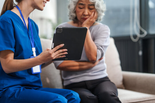 Caucasian Female Doctor Conducts A Check-up Using A Tablet Device For An Elderly Asian Patient Seated Together On The Sofa.