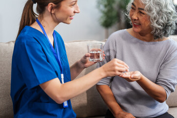 Caucasian female doctor provides a glass of water and medication pills to an elderly Asian patient while they are seated together on the sofa.