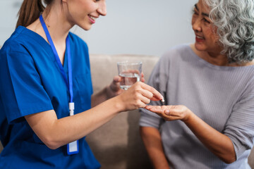 Caucasian female doctor provides a glass of water and medication pills to an elderly Asian patient while they are seated together on the sofa.