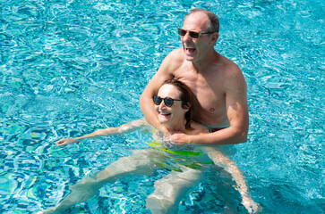 Bonding cheerful mature caucasian couple having fun floating in outdoor swimming pool under the sun. Vacation, healthy lifestyle and relax concept