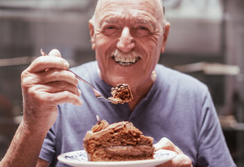 Portrait of senior man smiling while enjoying a break with a slice of chocolate cake. Greedy and...