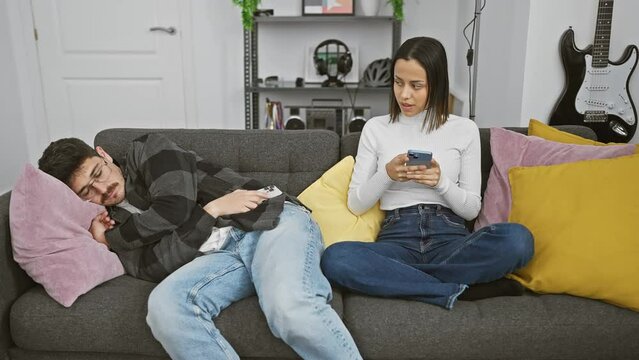In a modern living room, a woman checks a phone while a sleeping man lies oblivious next to her on the sofa.