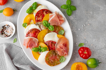 Salad of sliced multi-colored tomatoes, fresh peaches, mozzarella, prosciutto and basil on a gray concrete background. Delicious summer salad.