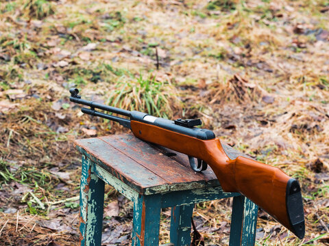 Classic airsoft or bb rifle with wooden stock and base and simple metal iron sight on old stool. Country side farm shooting range practice. Outdoor sport activity. Selective focus.
