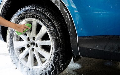 A person is washing a car tire with a green brush. Tire is covered in water and soap. Blue car wash with white soap foam. Auto care service. Car cleaning service concept. Vehicle cleaning service.