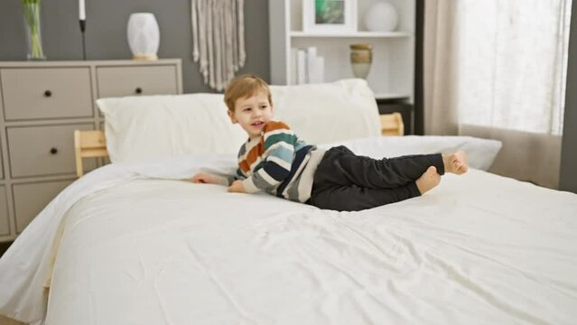 A playful boy in a striped shirt lies on a white bed in a cozy bedroom setting.
