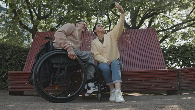 Cheerful man in wheelchair and his female friend taking selfie with mobile phone while spending day in park