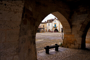 Monpazier Place des Cornières with an arch and a wooden bench in the foreground on a clear...