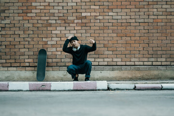 Fototapeta na wymiar Full body of Asian male in stylish clothes sitting with skateboard on sidewalk in the city