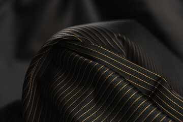 Close-up of texture of black lana wool fabric with pattern of thin stripes of gold color. Background, texture of draped suit fabric with shining pattern of gold-colored threads.