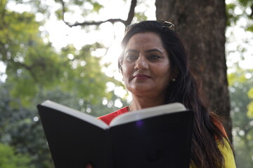 Portrait of mid aged woman holding a black color almanac in hand, reading personal almanac. sunlight, green park environment. book promotion, blank cover.