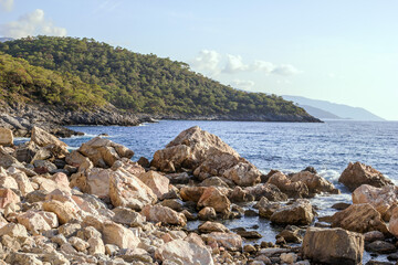 Beautiful small rocky beach with clear blue sea, waves and mountains on background. Oludeniz,...
