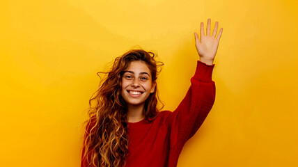 Young woman smiles and raises her hand in greeting on a yellow background. - 758764200