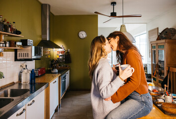Young female couple kissing while sharing a cup of tea in a in their home kitchen. Lesbian couple...