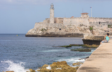 People sitting on the Malecon seawall of havana with a background view  of the fortress called The...