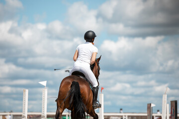 Young woman riding a horse. Close-up, view from the back. Competition on a sunny day, against a blue sky with clouds.