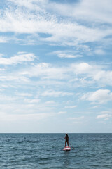 A woman in a hat on a SUP board in the sea against the background of the sky and clouds. Sunny day. Vertical.