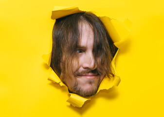 A long-haired, shaggy man with a beard and mustache looks out from a torn hole in yellow paper with...