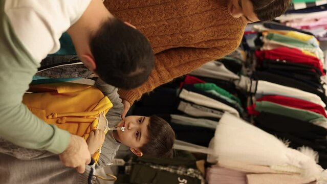 family shopping for clothes, mother, father and kids.children choosing in store, mother putting clothes in front of boy, father resting on sofa with phone and toddler playing with toys.4k