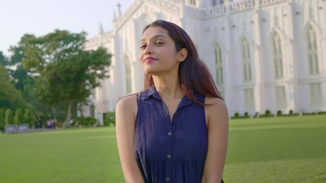 A young happy Indian Asian attractive woman or female student standing outdoors in front of the college or University building with a smile on her face in the daylight. Education, study abroad concept