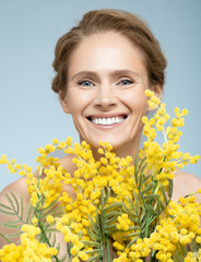 Skin and health care for women, concept. Beauty portrait of mature woman exuding vitality, clutching mimosa bouquet against blue background