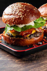 Close-up of a juicy burger with fresh lettuce, tomato, cheese, and a grilled patty on a sesame seed bun, served on a black plate. The background is dark, highlighting the vibrant colors of the burger