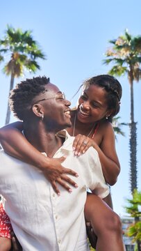 Vertical photo of a young African couple in love having fun together outdoors. Boyfriend giving piggyback to his girlfriend during summer vacation on a beach country.