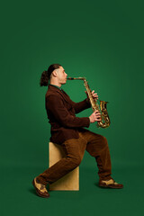 Talented and artistic young man, musician in costume sitting and playing saxophone against dark...