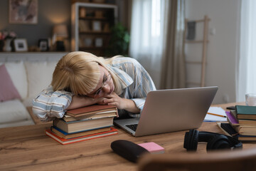 Young tired university student tired from study fro exam sleeping on the pile of books on desk. Exhausted woman leaning head on hand after mental burnout from over thinking