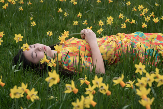 asian girl in yellow dress lying between daffodil flowers in field in spring