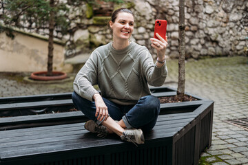 Obraz premium Happy 30s Women taking selfie and sitting on the bench outdoors. Young beautiful girl say Hi. Urban lifestyle concept, full body. High quality photo