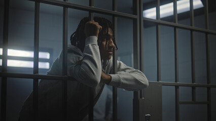 Upset African American teenager with face tattoos stands in prison cell in jail or youth detention center leaning on metal bars. Prison officer passes by young criminal or prisoner in the foreground. - Powered by Adobe