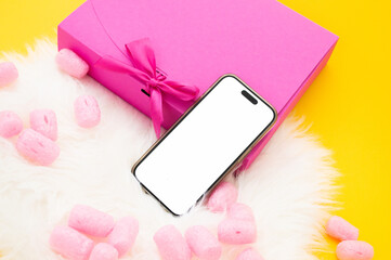 Bright and playful, the image presents a smartphone with a blank screen lying on a fluffy white surface, surrounded by pink marshmallows, with a vivid yellow background and a magenta gift box adorned 