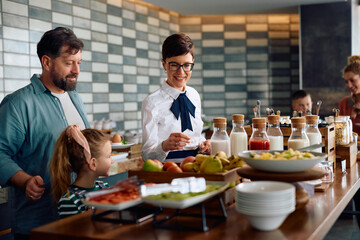 Happy waitress serving breakfast to family at buffet table in hotel.