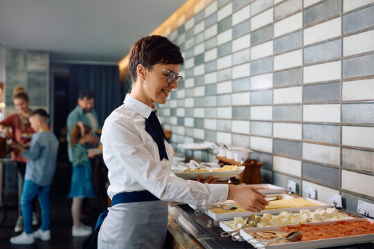 Happy waitress setting up buffet table for breakfast in hotel restaurant.