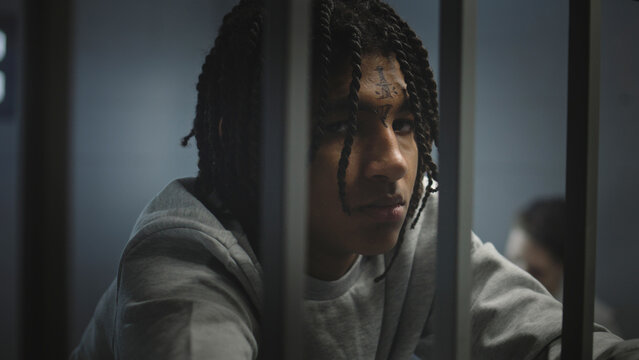 Angry African American teenage prisoner with face tattoos stands in prison cell in jail and looks at camera. Young criminal serves imprisonment term for crime. Juvenile detention center. Portrait.