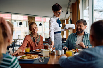 Happy waitress serving family during  breakfast in hotel restaurant. - obrazy, fototapety, plakaty