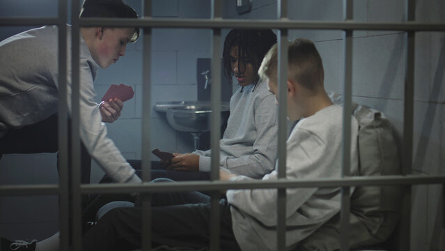 Three diverse teenagers play card games in prison cell. Young prisoners, criminals serve imprisonment term for crimes in jail. Youth detention center or correctional facility. View through metal bars.