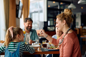 Happy mother feeding her daughter during family breakfast in hotel restaurant.