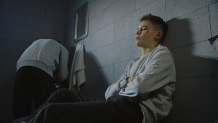 Upset Caucasian teenage prisoner sits on the bed in prison cell. African American inmate washes his...